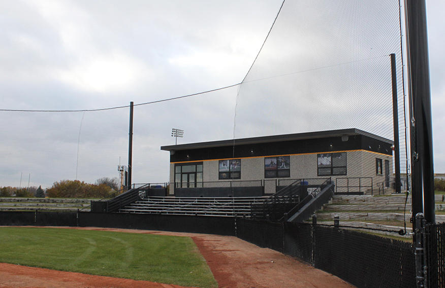 A baseball field with empty metal bleachers and a building behind home plate under a cloudy sky. Protective netting is stretched above the stands and grassy outfield is visible in the foreground.