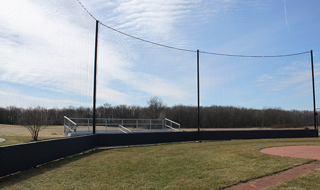 A baseball field with a grass outfield, a dirt infield, and tall netting behind the field, with empty bleachers in the background and trees under a partly cloudy sky.
