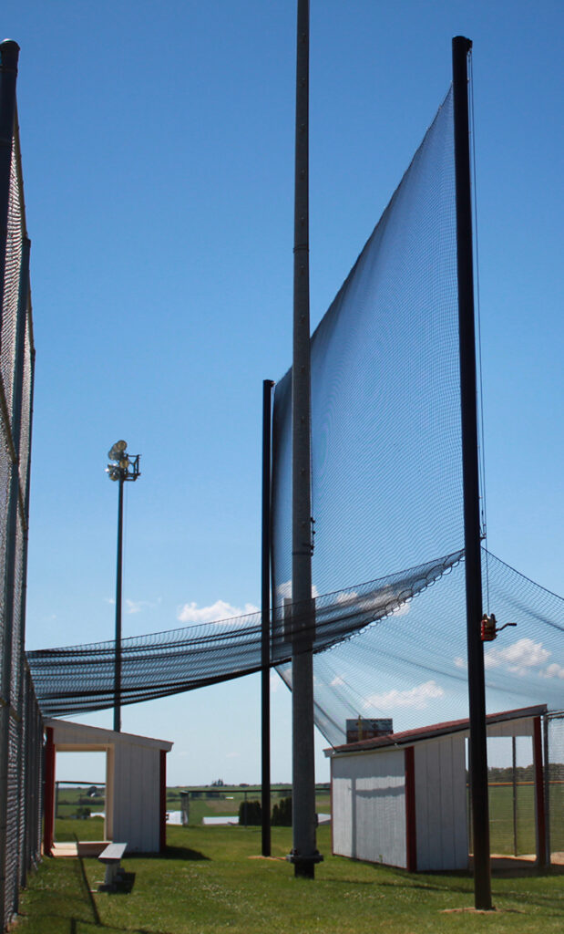 A tall safety net is suspended between poles near small white and red buildings at a baseball field, with a light post, fences, and green grass under a clear blue sky.