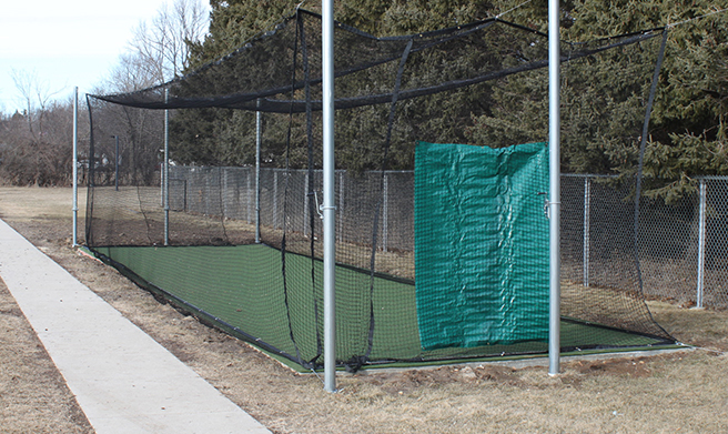 A batting cage with a green artificial turf floor and netting, surrounded by chain-link fencing and trees. A green tarp hangs on one side. A sidewalk runs along the left side of the cage.