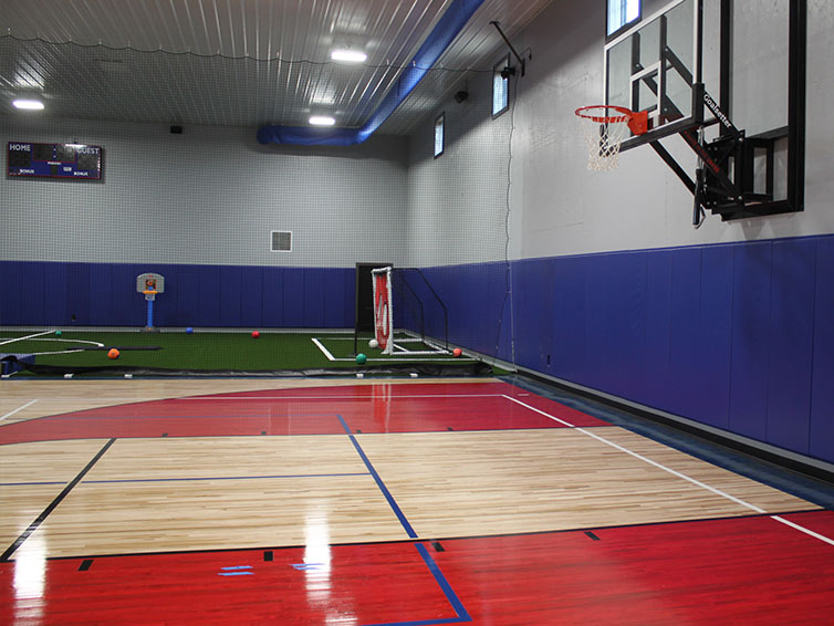 Indoor gymnasium with a basketball court in the foreground, a hoop on the right wall, and artificial turf area with soccer goals and various sports equipment in the background. Blue and gray walls surround the space.