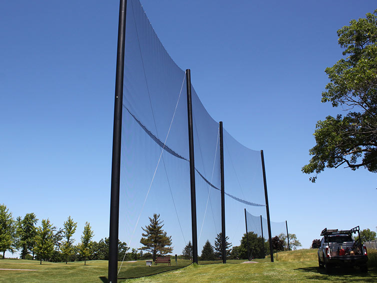Tall black poles supporting netting on a grassy area, likely a golf course driving range, with trees in the background and a blue sky above. A utility vehicle is parked on the right side.