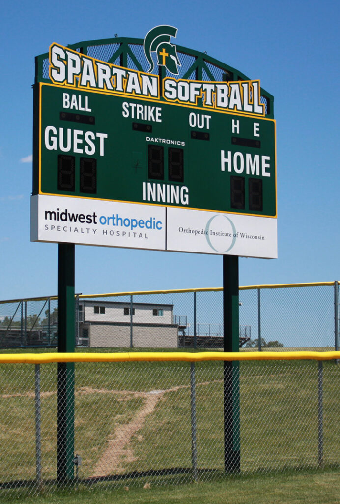 A green Spartan Softball scoreboard stands above a field with a yellow fence. The scoreboard shows spaces for ball, strike, out, inning, and scores for guest and home teams. A path and a building are visible in the background.