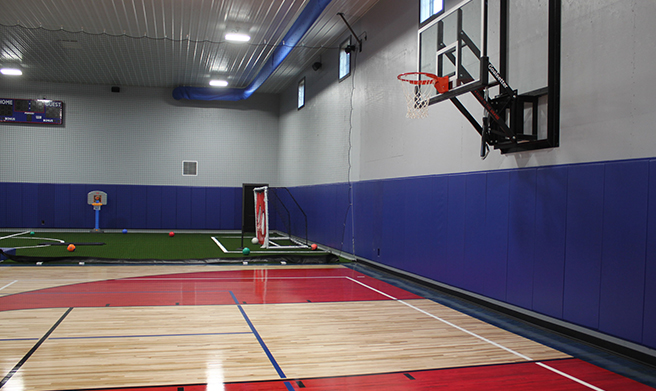 Indoor gym with a basketball hoop and court in the foreground; in the background, there is artificial turf with a small soccer goal, net, and various sports equipment scattered around. Walls are gray and blue.