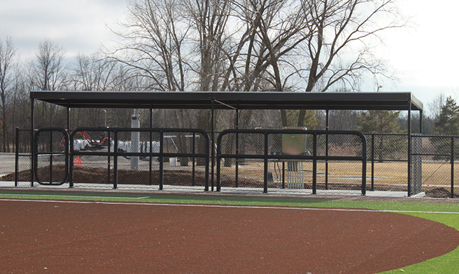 A fenced baseball dugout with a black metal roof stands next to a baseball field with brown turf. Leafless trees and cloudy skies are visible in the background.