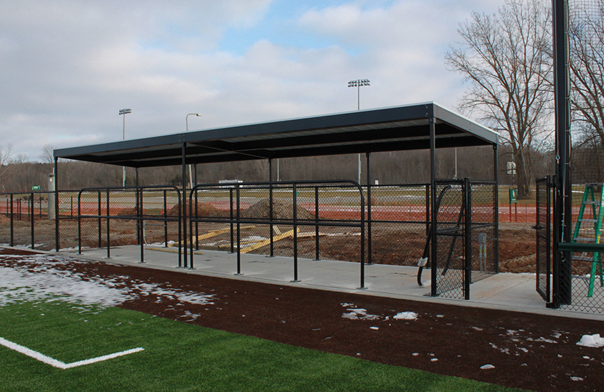 A covered baseball dugout with a chain-link fence sits beside an artificial turf field. In the background are trees, light poles, and other athletic fields under a cloudy sky. Some snow is visible on the ground.