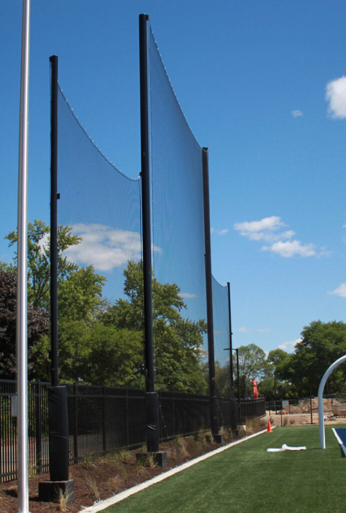 Tall black poles support a protective net behind a sports field, bordered by a fence and trees, under a clear blue sky with a few clouds.