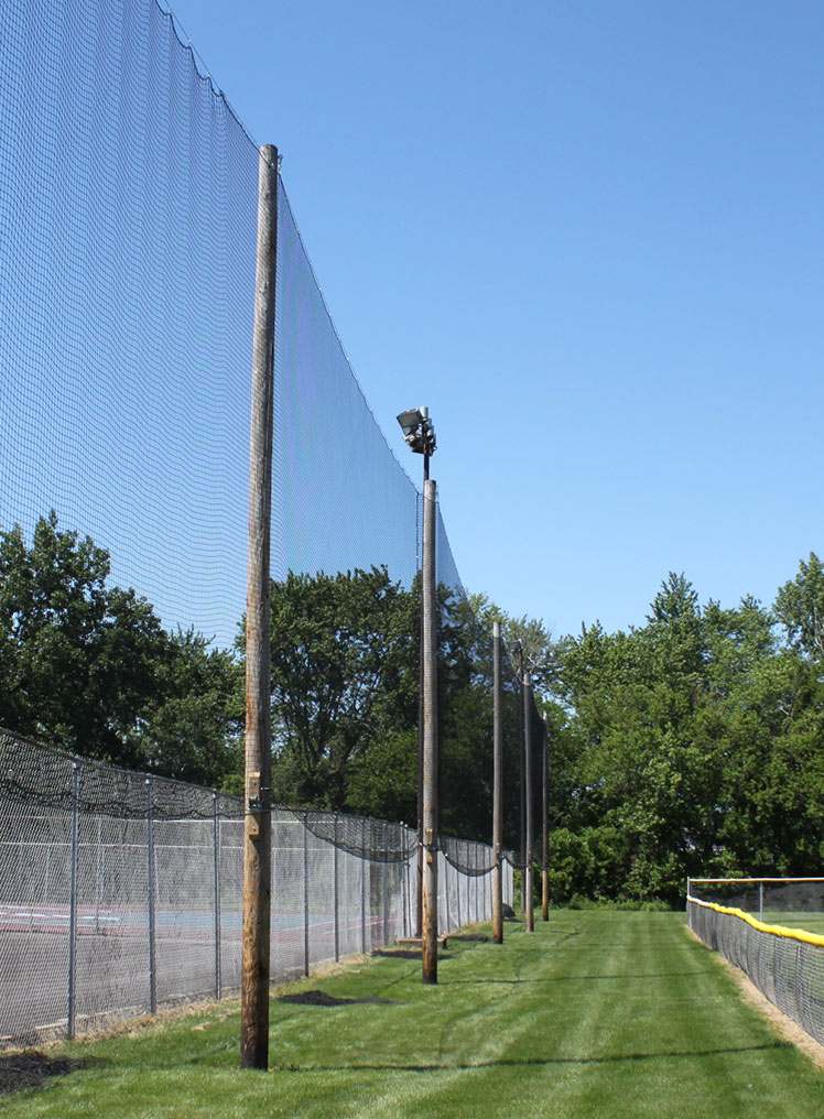 Tall wooden poles support a high protective net above a grassy area with chain-link fences and trees in the background under a clear blue sky.