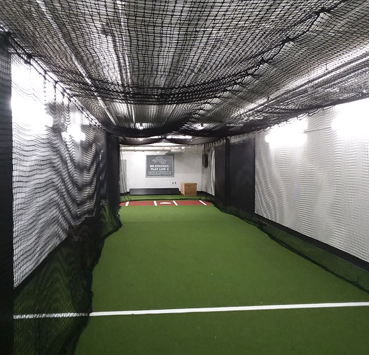An indoor batting cage with green artificial turf, netted walls and ceiling, white walls, and a pitching mound at the far end. Bright lights line the ceiling and a sign is visible on the back wall.