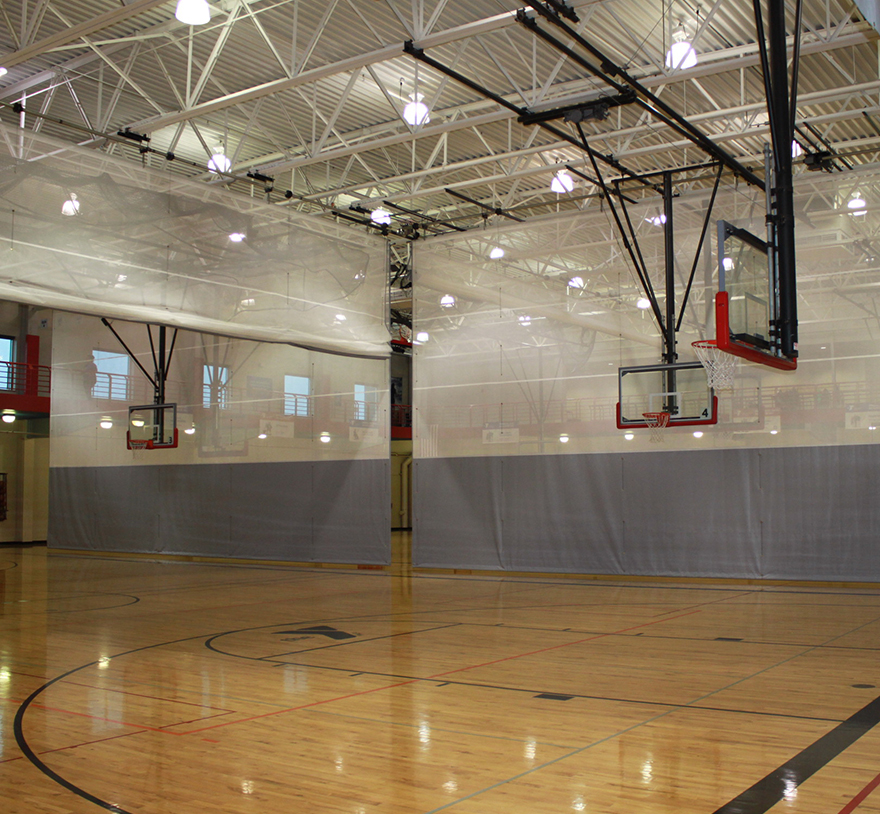An empty indoor basketball court with wooden flooring, two basketball hoops, high ceilings, and transparent divider curtains hanging down, separating the gym space. Bright overhead lights illuminate the area.